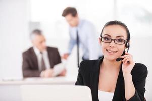 How may I help you Beautiful young woman in formalwear adjusting her headset and smiling while people working on background photo