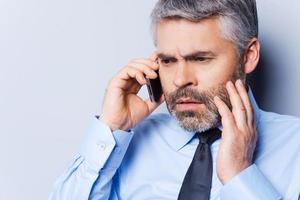 Bad news. Depressed mature man in shirt and tie talking on the mobile phone and touching face with hand while standing against grey background photo