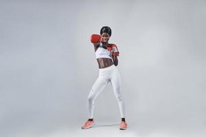 Beautiful young African woman in boxing gloves exercising while standing against gray background photo