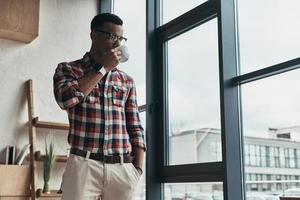On a little break. Handsome young man drinking coffee and keeping hand in pocket while standing near the window photo