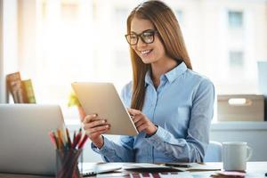 Lets check my timetable Cheerful young beautiful woman in glasses using her touchpad with smile while sitting at her working place photo