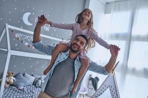 Carefree time with dad. Cheerful father carrying his daughter on shoulders and smiling while spending free time at home photo