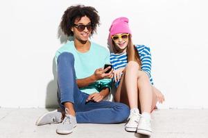 sin notar a nadie más. pareja joven funky sonriendo y mirando el teléfono móvil mientras se sienta contra el fondo blanco foto