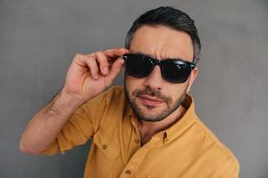 Well hello Top view of handsome young man adjusting his sunglasses and looking at camera while standing against grey background photo