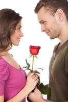 primera fecha. alegre joven pareja amorosa sosteniendo una rosa roja y sonriendo mientras está de pie cara a cara y aislado sobre fondo blanco. foto