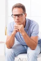 Confident expert. Confident casual businessman in glasses holding his hands on chin and looking at camera while sitting on the chair photo