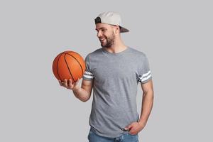 Ready to compete. Handsome young smiling man carrying a basketball ball and looking at it while standing against grey background photo