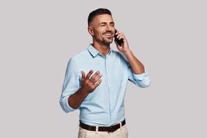 Good talk with client. Good looking young man in smart casual wear talking on the phone and smiling while standing against grey background photo