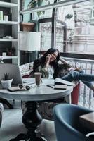 Having a little break. Attractive young smiling woman talking on smart phone and looking away while sitting in restaurant near the window photo