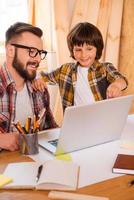 Working together with father. Happy young man working on laptop while his little son pointing at it photo