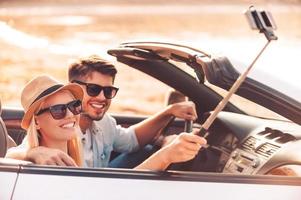 Selfie to remember the day. Happy young couple using monopod while making selfie inside of their convertible photo