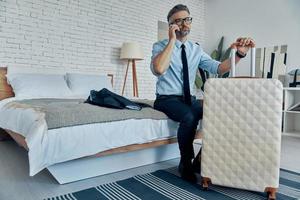 Handsome man in shirt and tie leaning on suitcase ant talking on phone while sitting on the bed photo