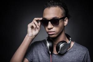 This is my style. Portrait of young African man adjusting his sunglasses and wearing headphones while standing against black background photo