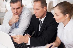 Business people at work. Three confident business people in formalwear sitting at the table and looking at the laptop while one of them pointing monitor photo