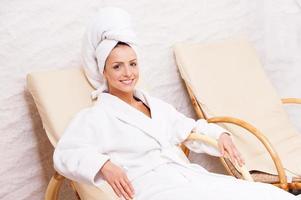 mujer en centro de spa. mujer joven atractiva en bata de baño y toalla en la cabeza relajándose mientras se sienta en una silla y sonríe foto