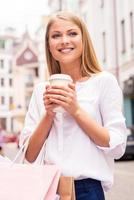 refrescarse antes de la próxima tienda. hermosa joven sonriente sosteniendo bolsas de compras y una taza de bebida caliente mientras está de pie al aire libre foto