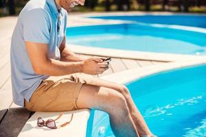 Relaxing by the pool. Close-up of young man in polo shirt sitting poolside and typing something on mobile phone photo