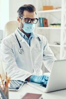 joven concentrado con bata de laboratorio blanca y máscara protectora que trabaja con una laptop mientras está sentado en el interior foto