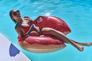 Let the worries float away. Attractive young woman in swimwear smiling while floating on the inflatable ring in the pool photo