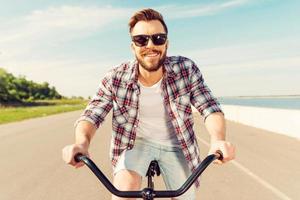la vida es más brillante cuando estás en bicicleta. un joven apuesto con anteojos sonriendo a la cámara mientras monta en bicicleta por la carretera foto