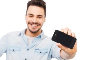 Look at my new gadget Cheerful young man showing mobile phone and smiling while standing against white background photo