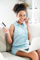 Enjoying online shopping. Attractive young African woman talking on the mobile phone and holding credit card while sitting on the couch with laptop at her knees photo