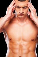 Awful headache. Young shirtless man touching head with hands and keeping eyes closed while standing against black background photo