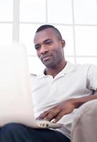 trabajando en casa. apuesto joven africano usando computadora y mientras está sentado en la silla foto