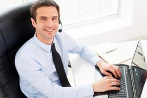 IT support. Top view of cheerful young man in headset looking at camera and smiling while using laptop photo