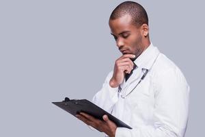 Thinking about proper medication. Thoughtful African doctor holding clipboard and looking at it while standing against grey background photo