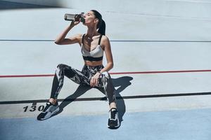 tan sediento. vista superior de una mujer joven con ropa deportiva bebiendo agua mientras se sienta en la pista de atletismo al aire libre foto