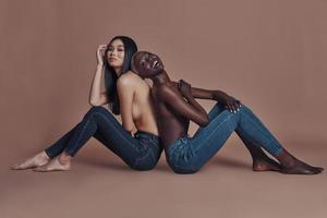 Supremely sensual. Attractive young topless women looking at camera and smiling while sitting back to back against brown background photo