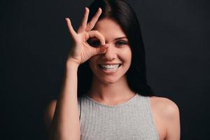 solo por diversión. hermosa mujer joven haciendo un gesto de ok cerca de su ojo y sonriendo mientras está de pie contra el fondo gris foto