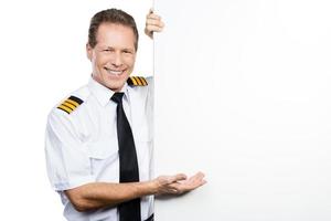 Can you believe it Confident male pilot in uniform leaning at the copy space and pointing it with smile while standing against white background photo