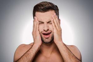 This awful headache Portrait of frustrated young shirtless man touching head with hands and keeping eyes closed while standing against grey background photo