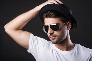 Confident in his perfect style. Handsome young man adjusting his hat and looking away while standing against black background photo