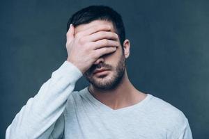 Bang Frustrated young man gesturing finger gun near head and keeping eyes closed while standing against grey background photo