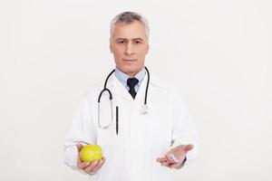 haga su elección médico senior de cabello gris en uniforme sosteniendo una manzana en una mano y pastillas en otra mientras está de pie aislado en blanco foto