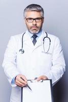 Copy space on his clipboard. Confident mature doctor looking at camera and pointing copy space on his clipboard  while standing isolated on white photo