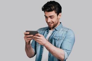 Feeling playful. Good looking young man in blue jeans shirt using his smart phone while standing against grey background photo