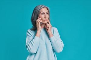 hermosa mujer madura hablando por teléfono inteligente mientras está de pie contra el fondo azul foto