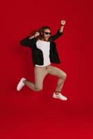 Full length of cheerful young man in casual clothing smiling and gesturing while hovering against red background photo