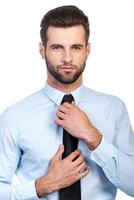 Used to look perfect. Confident young handsome man adjusting his necktie and looking at camera while standing against white background photo