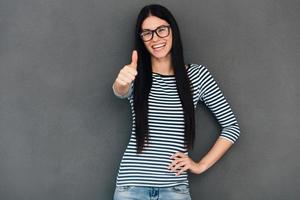 Everything is alright  Attractive young woman showing her thumb up and looking at camera with smile while standing against grey background photo