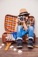 Catching the best moments. Little boy in headwear holding camera and smiling while sitting in suitcase against brown background photo