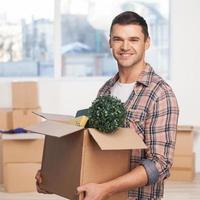 Just moved in a new apartment. Cheerful young man holding an opened cardboard box and smiling at camera while more carton boxes laying on background photo