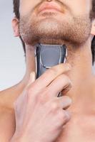 Man shaving. Close up of young man shaving his face with electric shaver while standing against grey background photo