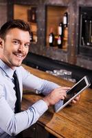 navegando por la red en el bar. un joven apuesto con camisa y corbata trabajando en una tableta digital y sonriendo mientras se sienta en el mostrador del bar foto