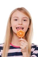 Girl with lollipop. Cheerful little girl licking lollipops and looking at camera while standing isolated on white photo