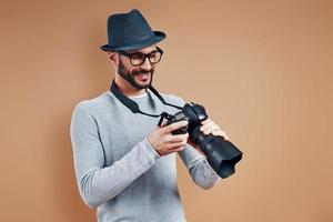 un joven apuesto con ropa informal sonriendo y ajustando la cámara fotográfica mientras se enfrenta a un fondo marrón foto